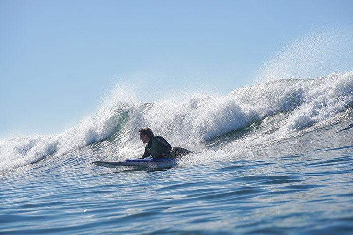 Eine Welle. Vor der Welle ein Surfer halbliegend auf einem Surfbrett im Wasser. Er schaut nach rechts. Wolkenloser hellblauer Himmel.