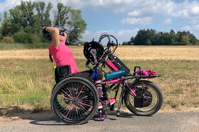 Eine junge Frau mit ihrem schwarz-pinken Handbike unterwegs in der Natur. Sie trägt einen Fahrradhelm und hält sich mit den Armen am Hinterteil des Helms fest.