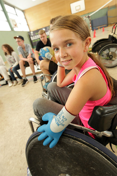 Ein junges Mädchen in einem Rollstuhl. Sie lächelt. Mit ihrem rechten Arm stützt sie sich auf ihrem rechten Bein ab. An der linken Hand trägt sie einen Handschuh.