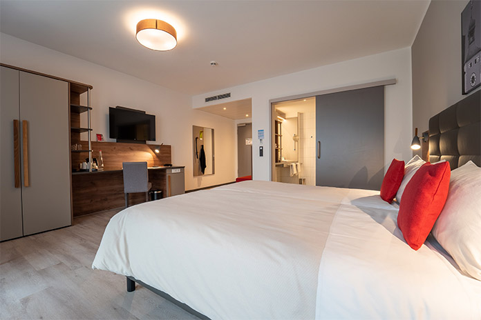 Ein Hotelzimmer ausgestattet mit einem großen Doppelbett, einer Schiebetür zum Bad, einem Schrank, Ablagefläche und Stuhl, TV und einem Spiegel. 
