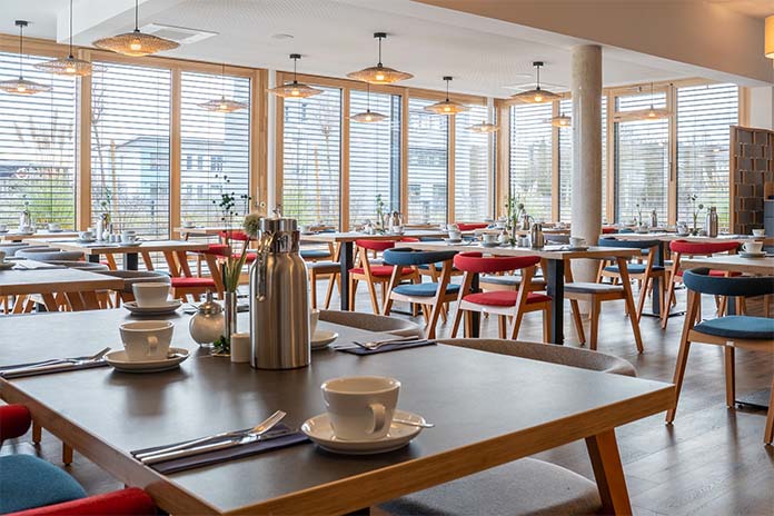 Ein Restaurant in einem Hotel mit einzelnen Tischen und Stühlen. Das Polster der Stühle ist in den Farben rot und blau. Die Tische sind mit Kaffeetassen und Kanne, Messern und Gabeln eingedeckt. An den Fenstern befinden sich Jalousien.