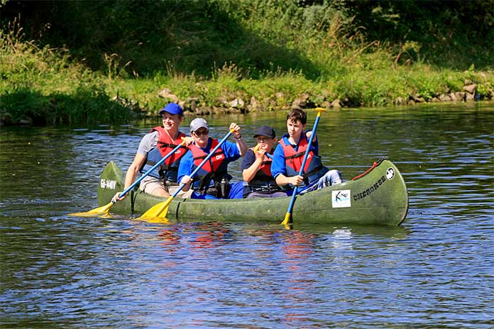 Ein Kanu auf einem Gewässer mit vier Jugendlichen. Der Junge ganz vorne im Kanu hält sein Paddel mit der rechten Hand. Sein linker Unterarm ist verkürzt. Der zweite Junge hält das Paddel auf der linken Seite. Die beiden hinteren Jungs halten das Paddel auf der rechten Seite. 