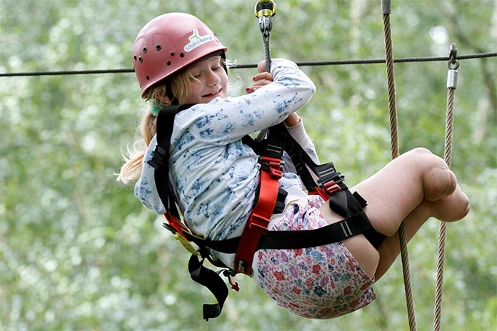 Ein Mädchen in einem Klettergarten. Sie hängt angeschnallt mit einem Sicherheitshelm an einem Seil. Ihre Beine sind verkürzt. Ihr fehlen beide Unterschenkel und Füße.