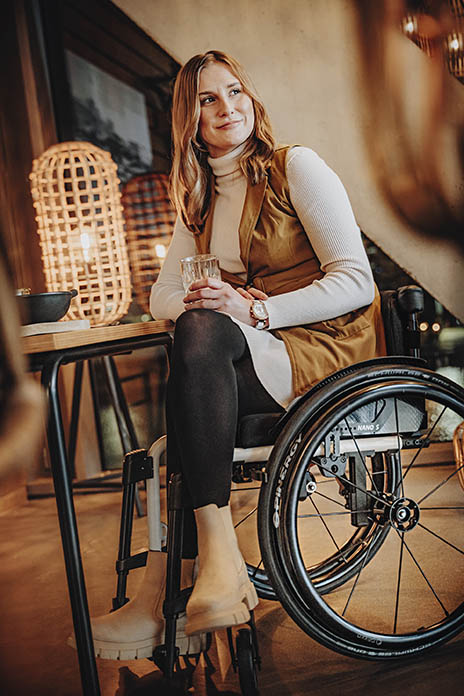 Eine junge Frau lächelnd in einem Rollstuhl. Sie befindet sich in einem Café. In ihren Händen hält sie ein Glas, abgestützt auf ihrem Bein. Die Beine liegen übereinander.