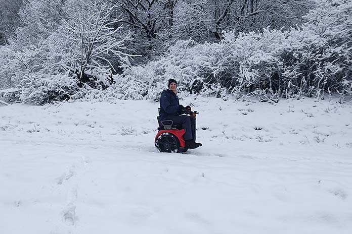 Eine Frau in einem roten, sportlichen Segway Rollstuhl fahrend durch eine Schneelandschaft. Sie hält sich an einer Lenkstange fest. 