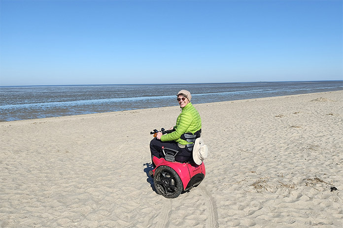 Eine Frau in einem roten, sportlichen Segway Rollstuhl stehend am Sandstrand. Vor ihr ist das Meer. An ihrem Rollstuhl befindet sich ein Sonnenhut. Sie selbst trägt eine grüne Jacke. 