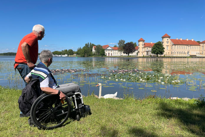 Ein älterer Herr und eine ältere Dame im Rollstuhl, beide stehen vor einem See. Im Hintergrund das Schloss Sanssouci.