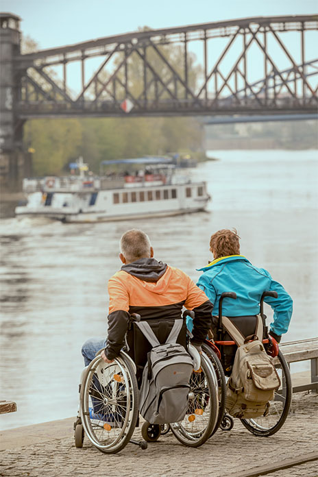 Zwei Rollstuhlfahrer an einem Ufer. Rechts eine Frau links ein Mann. An den Rollstühlen hängt jeweils ein Rucksack. Sie schauen auf ein Schiff. Über dem Schiff ist eine Brücke.