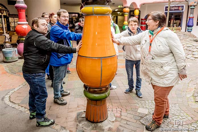 In einem Kreis stehen mehrere Menschen. Sie halten sich mit der rechten Hand an einem Gefäß fest. Es ist aus Keramik gefertigt, orangefarben und bauchförmig. Eine Frau auf der rechten Seite trägt ein Schild um den Hals. Sie führt die Gruppe.