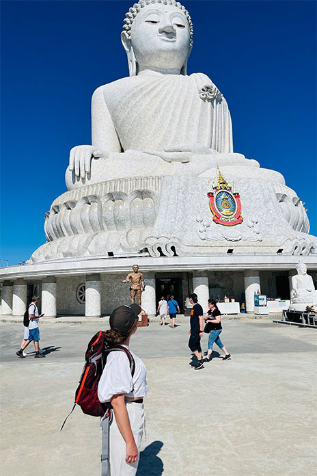 Ein helles Buddahdenkmal gestützt auf Säulen. Davor laufen ein Menschen. Der Himmel ist blau. Vorne im Bild eine junge Frau mit Rucksack und Cappy.