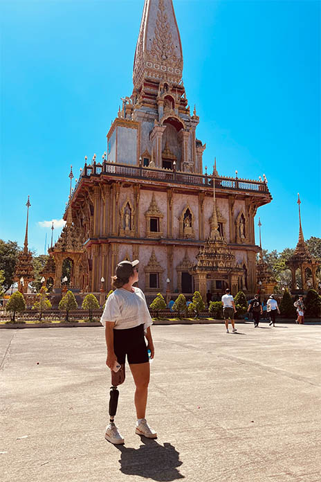 Ein größes, mehrstöckiges Denkmal in Thailand. Vorne im Bild eine junge Frau. Sie schaut seitlich auf das Denkmal. Trägt an ihrem rechten Bein eine Prothese, ein schwarzes Cappy und hält in ihrer rechten Hand ein Handy.