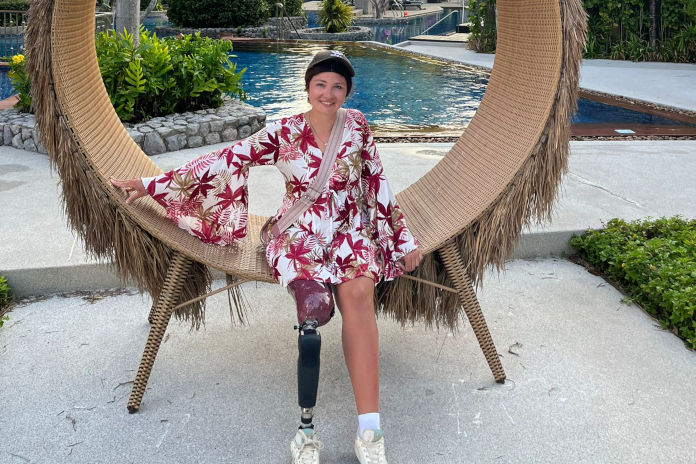 Eine junge Frau sitzend in einem Rattan Gestell mit Palmenwedeln. Am rechten Bein trägt sie eine Prothese. Im Hintergrund befindet sich ein Hotel.