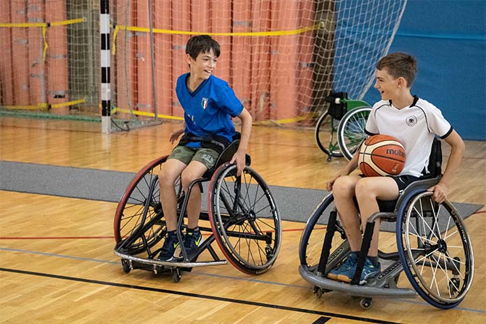 Zwei Jungs in Sportrollstühlen nebeneinander in einer Sporthalle. Beide schauen sich gegenseitig an und lachen. Beim rechten Jungen liegt ein Basketball auf den Beinen.