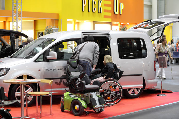 In einer Messehalle steht ein VW Caddy mit offener Heckklappe, ein Mann und eine Frau im Rollstuhl schauen sich das Auto an. Die Seitentür des Autos ist ebenfalls geöffnet.