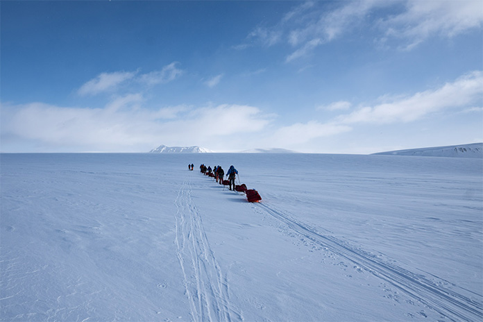 Eine Gruppe von Wanderern auf Skiern läuft in einer weitläufigen Schneelandschaft dem Horizont entgegen.
