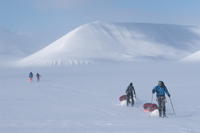 In einer weißen Schneelandschaft laufen zwei Menschen mit Skiern, im Hintergrund ein Schneeberg und zwei weitere Menschen.