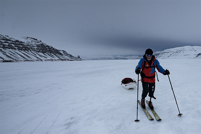 Ein Mann in einer Schneelandschaft umgeben von Bergen. Er steht auf Skiern. Am linken Bein trägt er eine Prothese. In den Händen hält er Skistöcker zur Fortbewegung. Hinter sich zieht er an einer Leine ein Boot.