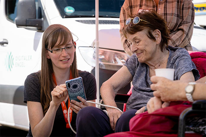Eine im Rollstuhlsitzende ältere Dame. Rechts neben ihr eine junge Frau. Sie hält in ihrer linken Hand ein Smartphone und hält ihren rechten Zeigefinger auf dieses. Die junge Frau erklärt etwas. Die ältere Dame schaut zu. Beide lächeln.