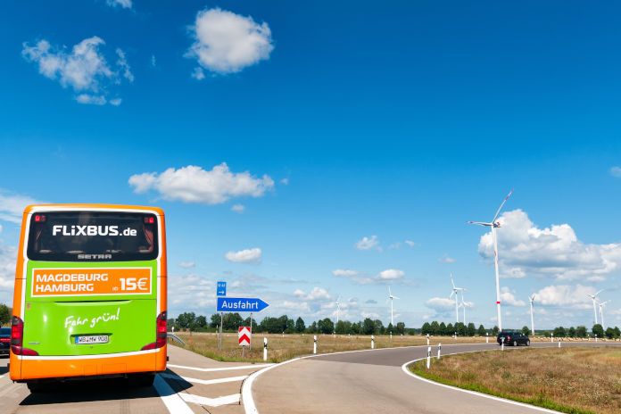 Eine grüner Flixbus fährt auf der Autobahn an einer Ausfahrt vorbei. Der Himmel ist blau. 
