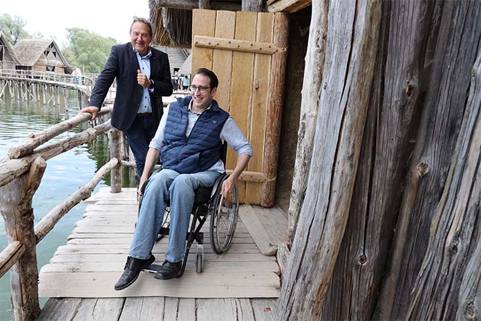 Auf einem Holzsteg an einer Holzhütte. Ein Mann im Rollstuhl lachend. Hinter ihm steht ein weiterer Mann ebenfalls lächelnd. Er hält sich an einem Holzgeländer fest. Unter dem Steg befindet sich Wasser.