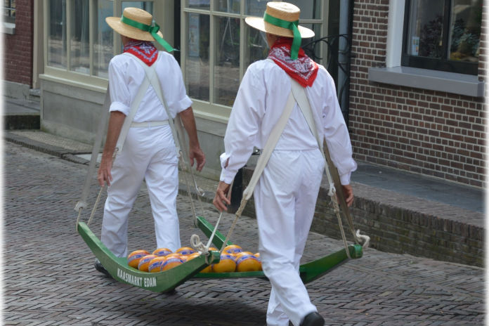 Zwei Männer in weißer Kleidung (Tracht) und Hut. Von hinten aufgenommen. Sie tragen auf einem grünen Holzgestell liegenden Edamer Käse.