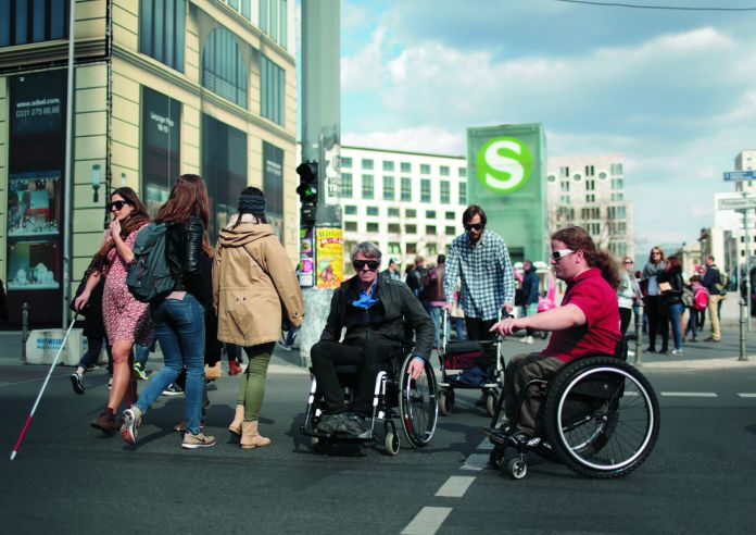 Beim überqueren einer Straße. Zwei Rollstuhlfahrer, ein Mann mit einem Rollator, eine Frau mit Blindenstock. Im Hintergrund eine Ampel, mehrere Gebäude und ein S-Bahn Hinweisschild.