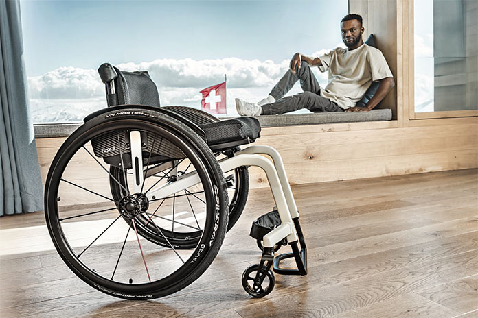 Im Vordergrund steht ein weißer Rollstuhl mit kantigem Rahmen. Im Hintergrund sitzt ein Mann auf einem Fensterbrett, dahinter Berge