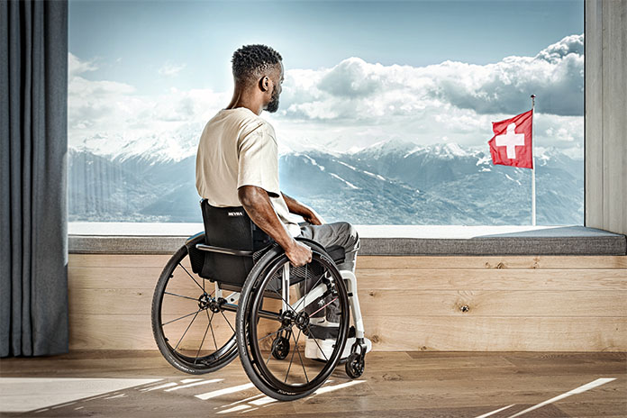 Ein Mann im Rollstuhl schaut aus einem großem Fenster, dahinter eine schweizer Flagge und Berge 