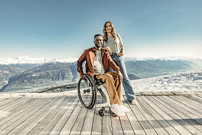 Ein Mann sitzt im Rollstuhl, neben ihn steht eine Frau, sie hat ihre Hand auf seiner Schulter. Sie stehen auf Holzfußboden, im Hintergrund ist ein Bergpanorama zu sehen