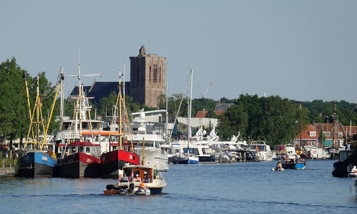 Ein historisches gebäude mit einem Turm, davor ein Hafen und Wasser. Ein Motorboot fährt aus dem Hafen heraus.