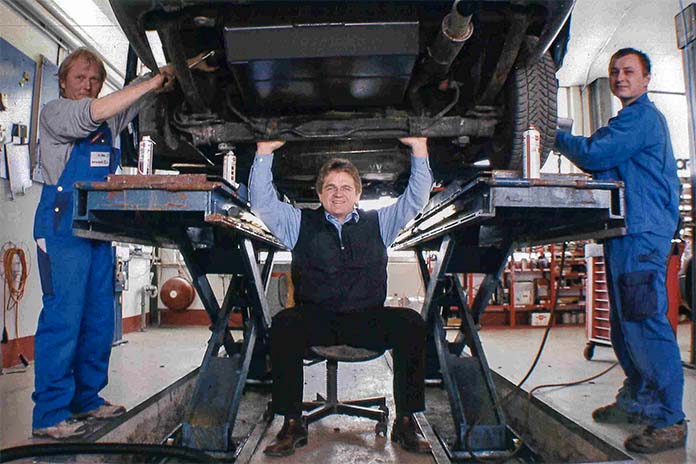 Ein altes Foto von einer Werkstatt in dem ein Mann, der unter einem auf der Bühne hochgefahrenen Auto sitzt und seine Arme nach oben unter die Achse hebt. 