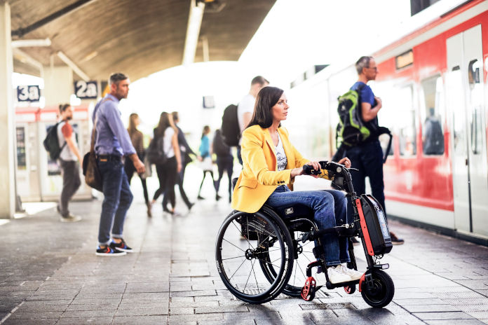 Eine Frau im Rollstuhl steht mit einem Zuggerät vor dem Rollstuhl auf einem Bahnhof. Ein Zug steht bereit.