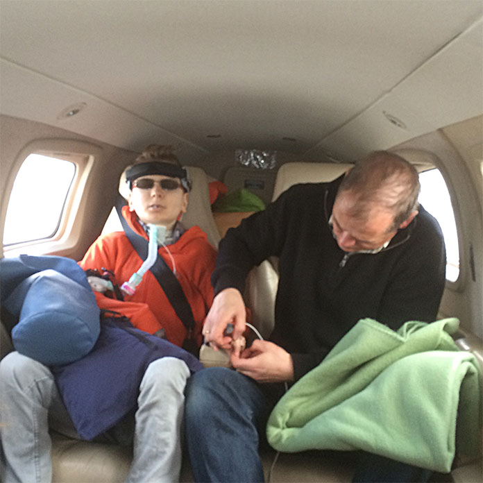 In einem Kleinflugzeug. Auf der linken Seite ein Patient, mit medizinischen Geräten. Auf der rechten Seite ein Mann. Er versorgt den Finger des Patienten. Auf seinem Bein liegt eine grüne Wolldecke. 