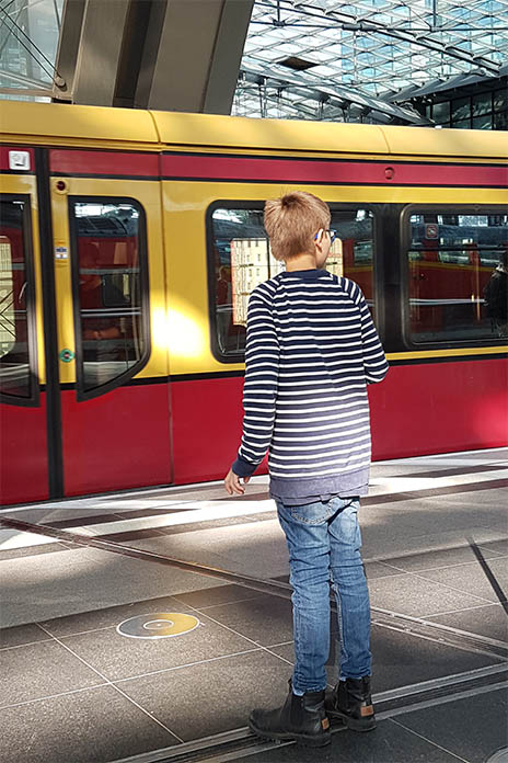 In einem Bahnhofsgebäude. Ein Junge steht am Bahngleis. Er schaut nach rechts auf einen rotgelben Zug, der vor im fährt. 