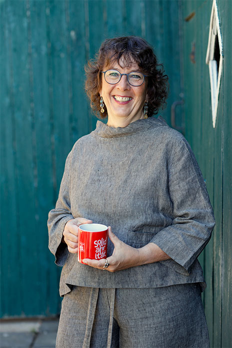 Eine Frau vor einer grünen Holzlaube, in einem grauen Leinen Zweiteiler gekleidet. Sie lacht. Trägt eine Brille und etwas längere Ohrringe. In den Händen hält sie einen roten Kaffeebecher. 