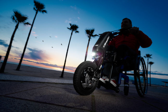 Ein Rollstuhlfahrer mit Zuggerät im Dunkel. Das Zuggerät hat Beleuchtung. Im Hintergrund Palmen. 