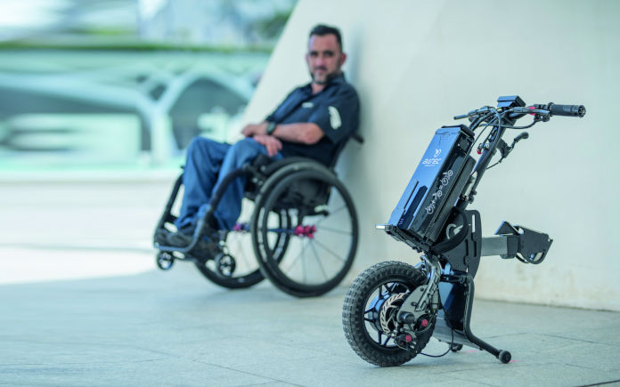 Im Vordergrund steht ein Zuggerät für Rollstühle, im Hintergrund lehnt ein Rollstuhlfahrer an einer grauen Betonwand.