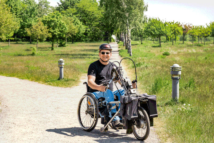 Ein Mann sitzend mit seinem Handbike auf einem Sandweg. Er lächelt, trägt eine Sonnenbrille und einen Fahrradhelm. Umgeben von Bäumen und Wiesen. Fotoquelle: BV_BFW_Kruppa_Daniel_S.gif 