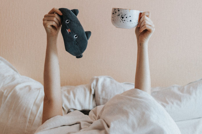 In einem Bett liegt eine Person. Die Bettdecke ist bis zum Kopf hochgezogen. Die Arme sind gestreckt. In der rechten Hand hält die Person eine Schlafmaske in Katzenform, in der linken eine große Kaffeetasse. Im Hintergrund befinden sich zwei Kissen.