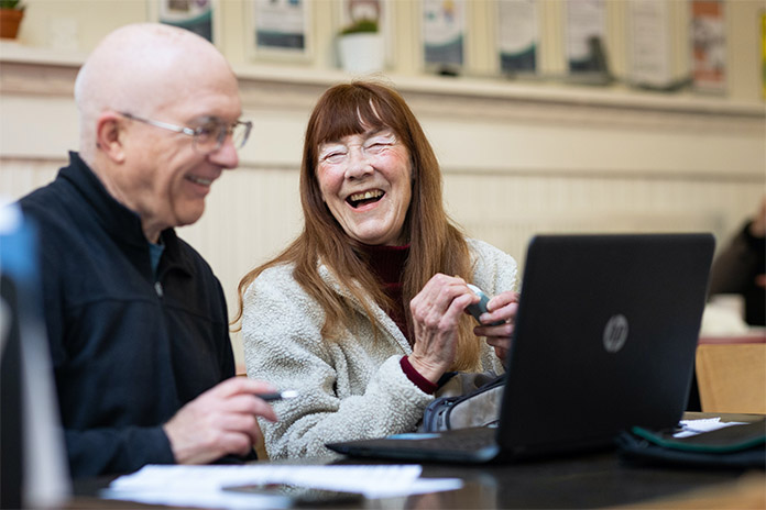 Ein älterer Mann links im Bild, neben ihm sitzt eine ältere Frau. Beide sitzen vor einem Laptop und lachen.