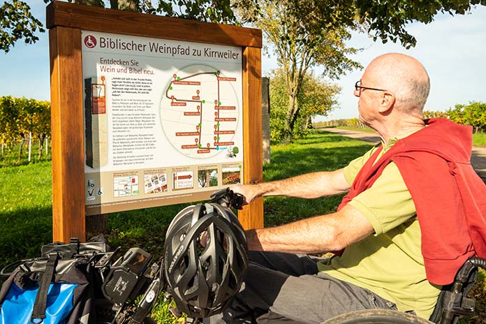 Eine Informationstafel aus Holz in der Umgebung eines Weinpfades. Davor ein Mann in seinem Handbike sitzend. An dem Lenker hängt sein Fahrradhelm. Er trägt einen Pullover über die Schultern