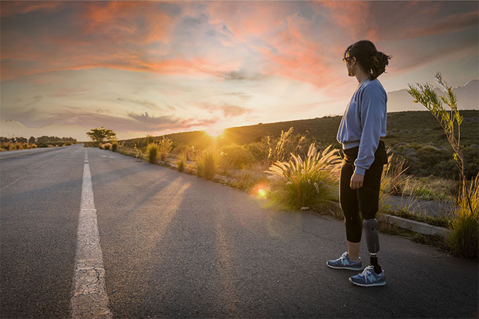 Eine Straße im Licht eines Sonnenunterganges. Rechts im Bild eine Frau. Sie trägt am linken Bein eine Unterschenkelprothese und schaut zur Sonne.