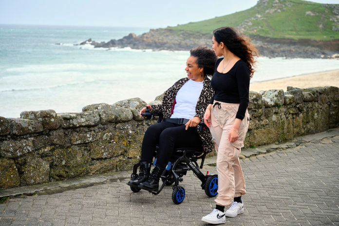 Auf einer Straße nahe der Küste geht eine jüngere Frau neben einer älteren in einem elektrischem Rollstuhl. Beide schauen auf das Meer hinaus.