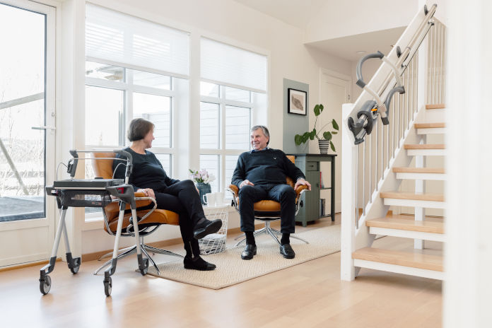 Zwei ältere Menschen sitzen in einem hellen Raum mit großen Fenstern. Ein Rollator steht neben der einen Person. An der Treppe ist der TOPRO Step angebracht. 
