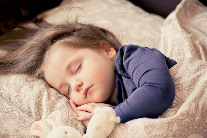 Ein schlafendes Kind auf einem Kopfkissen zugedeckt. Seine Hände hält es an der Wange. Daneben liegt ein Kuscheltier.