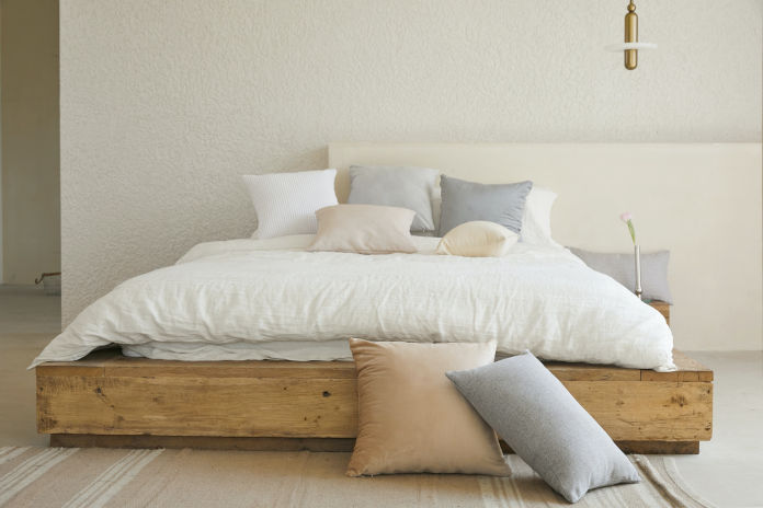 Ein Holzbett an einer Wand stehend. Drapiert mit verschiedenen Kissen und einer Bettdecke.