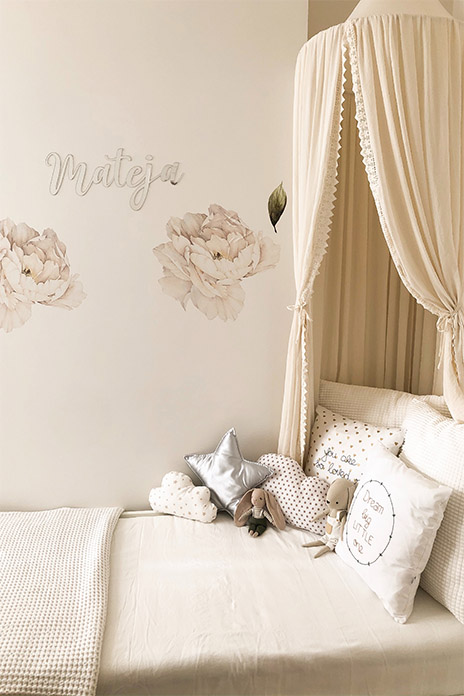 Ein Kinderbett umgeben mit verschiedenen Kopfkissen, Kuscheltieren, Tagesdecke und einem Betthimmel. An der Wand befinden sich zwei Wandtatoos in Rosenform. Alles in beigefarbenen, zarten Farbtönen.