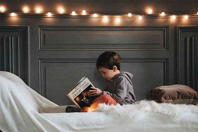 Ein kleiner Junge sitzt auf einem Bett und schaut sich ein Buch an. Hinter ihm hängt eine Lichterkette