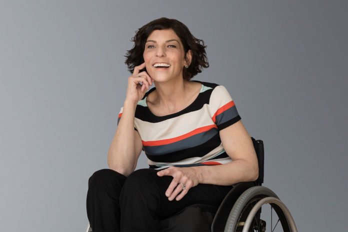 Eine Frau sitzend in einem Rollstuhl. Sie lacht und stützt ihren rechten Arm angewinkelt auf dem Bein ab.