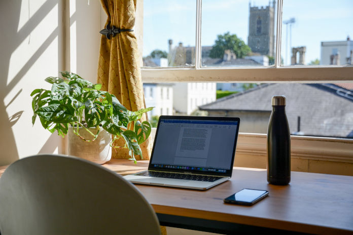 Ein Schreibtisch vor einem Fenster mit Ausblick. Auf dem Schreibtisch stehen ein Laptop, eine Thermoskanne und es liegt ein Handy.